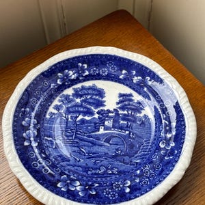 Może przedstawiać: Błękitno-biały talerz ceramiczny z błękitno-białym kwiatowym obramowaniem i błękitno-białym krajobrazem w środku. Scena przedstawia most, drzewa i dom.