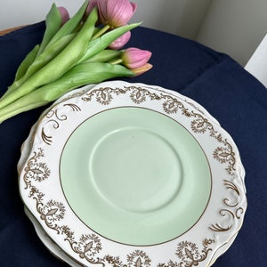 Puede incluir: Una pila de platos vintage con centro verde claro y borde blanco, adornados con filigrana dorada. Los platos tienen un borde festoneado. Un ramo de tulipanes rosas descansa junto a los platos sobre una superficie azul oscuro.