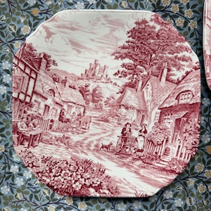 May include: An octagonal plate with a red and white village scene. The plate depicts a rural landscape with houses, a road, and a castle. The plate is set against a blue floral patterned background.