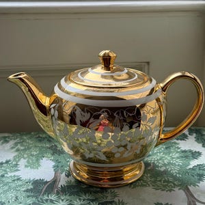 Könnte beinhalten: Eine goldfarbene Teekanne mit weißen floralen und gestreiften Akzenten. Die Teekanne hat einen gebogenen Griff, einen Ausguss und einen Deckel mit einem kleinen Knopf. Die Teekanne steht auf einem Sockel. Die Teekanne dient wahrscheinlich zum Servieren von Tee.