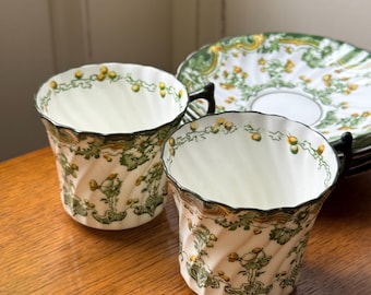 May include: Two white teacups with a green and yellow floral design and black handles. The cups have a twisted pattern and are accompanied by matching saucers with a similar design. The set is on a wooden surface.