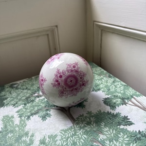 May include: A spherical ceramic object with a white base and a crackled glaze finish, decorated with repeating magenta floral patterns. The surface features intricate clusters of small flowers and larger central blooms arranged symmetrically.