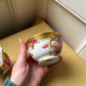 Op de afbeelding: Een witte porseleinen theekop met een gouden rand en een bloemenrozenpatroon. De theekop wordt in een hand gehouden, waarbij het delicate ontwerp wordt benadrukt. Een klassiek stuk voor theepartijen of presentatie.