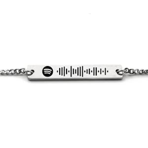 Puede incluir: Un brazalete de cadena de plata con un dije rectangular que presenta un logotipo de Spotify y un código de barras.