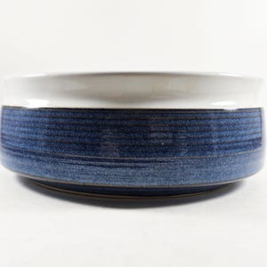 May include: A round ceramic bowl featuring a smooth white interior and a textured exterior with horizontal bands of deep blue and navy glaze. The bowl has a wide, slightly flared rim and a low profile, showcasing a distinct contrast between the white top and the dark blue base.