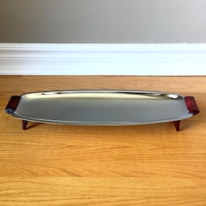 Peut inclure: Un plateau de service oblong vintage avec une surface polie argentée. Le plateau a des poignées rectangulaires rouge foncé à chaque extrémité et est soutenu par quatre petits pieds triangulaires rouges. Le plateau est présenté sur une surface en bois.