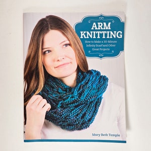 May include: A book cover titled "ARM KNITTING" featuring a woman wearing a blue infinity scarf. The book promises instructions on how to make a 30-minute infinity scarf and other projects. The author's name, Mary Beth Temple, is also visible.