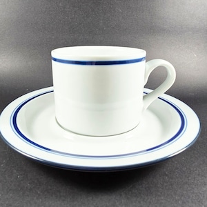 May include: White ceramic teacup and saucer set. The cup has a cylindrical shape with a handle and a blue stripe around the rim. The saucer also has a blue stripe around the edge. Ideal for serving tea or coffee.