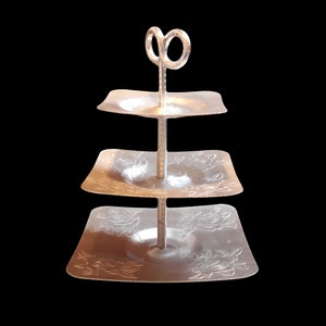 May include: A three-tiered silver-coloured metal serving stand with square trays. The two lower trays feature embossed rose floral patterns. A central vertical rod connects the tiers, topped with a circular handle loop for carrying the stand.