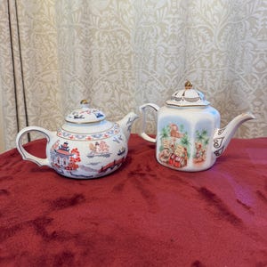 Op de afbeelding: Twee witte porseleinen theepotten met gouden randen. De linker theepot heeft een ronde vorm met een rood en blauw pagodelandschap. De rechter theepot heeft een zeshoekig lichaam versierd met een kleurrijke scène met figuren.