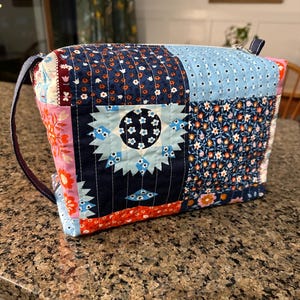 May include: A patchwork zippered bag with a navy blue strap. The bag is made of various fabric squares in shades of blue, pink, and red, with floral and geometric patterns. The bag has a rectangular shape and a zipper closure.