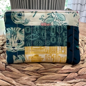 May include: A small, quilted coin purse with a zipper closure. The patchwork design features cat faces, floral patterns, and keyhole motifs in teal, cream, and yellow. The purse is resting on a woven mat.