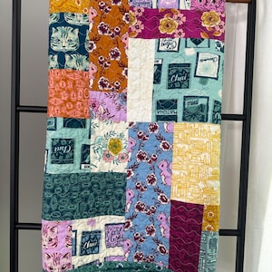 May include: A colorful patchwork quilt with a variety of patterns, including cats, flowers, and books. The quilt is made up of squares of different fabrics, each with a unique design. The quilt is hanging on a black rack.