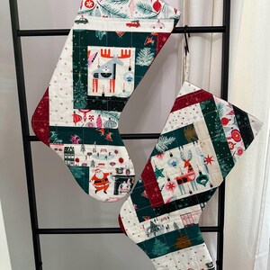 May include: Two Christmas stockings, one with a moose design and the other with a reindeer design. Both stockings are made of patchwork fabric with a white background and green, red, and white patterns. The stockings are hanging on a black metal rack. The text "Option A - Moose" is above the moose stocking and the text "Option B - Reindeer" is below the reindeer stocking.
