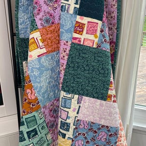May include: A patchwork quilt with a variety of patterned fabrics in shades of blue, pink, green, and orange. The quilt features floral, cat, and tea-themed designs. The quilt is draped over a black metal rack.