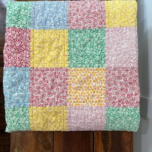May include: A patchwork quilt with a variety of floral patterns in squares. The quilt features squares of red, green, yellow, blue, and pink floral fabrics. The quilt is folded on a wooden surface.