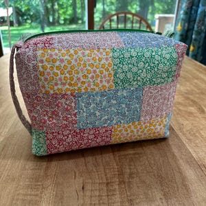 May include: A rectangular zippered pouch constructed from quilted fabric featuring a patchwork design of various floral patterns in shades of red, yellow, green, and blue. The bag includes a green zipper closure and a matching fabric side handle.
