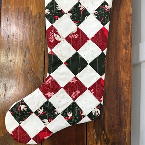 May include: A quilted Christmas stocking with a red hanging loop. The stocking features a patchwork design of red, white, and dark green diamond shapes. Some diamonds have floral and bird patterns. The stocking is hanging on a wooden surface.