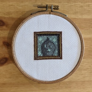 Puede incluir: Un aro de bordado blanco con un marco de madera marrón. El aro contiene un diseño de punto de cruz de una imagen azul y verde enmarcada en marrón.