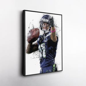 May include: A framed art print of a football player in a navy blue uniform, holding a football and giving a thumbs-up. The artwork has a watercolor effect with paint splatters and a white background. The player wears a helmet and a pink wristband.