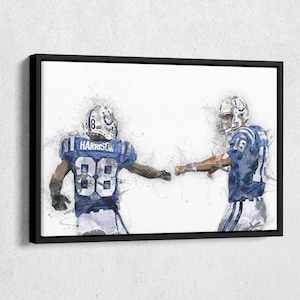 May include: Framed artwork featuring two football players in blue and white uniforms, with the number 88 and the name "Harrison" visible on one jersey. The players are fist-bumping, set against a white background with artistic paint splatter effects.