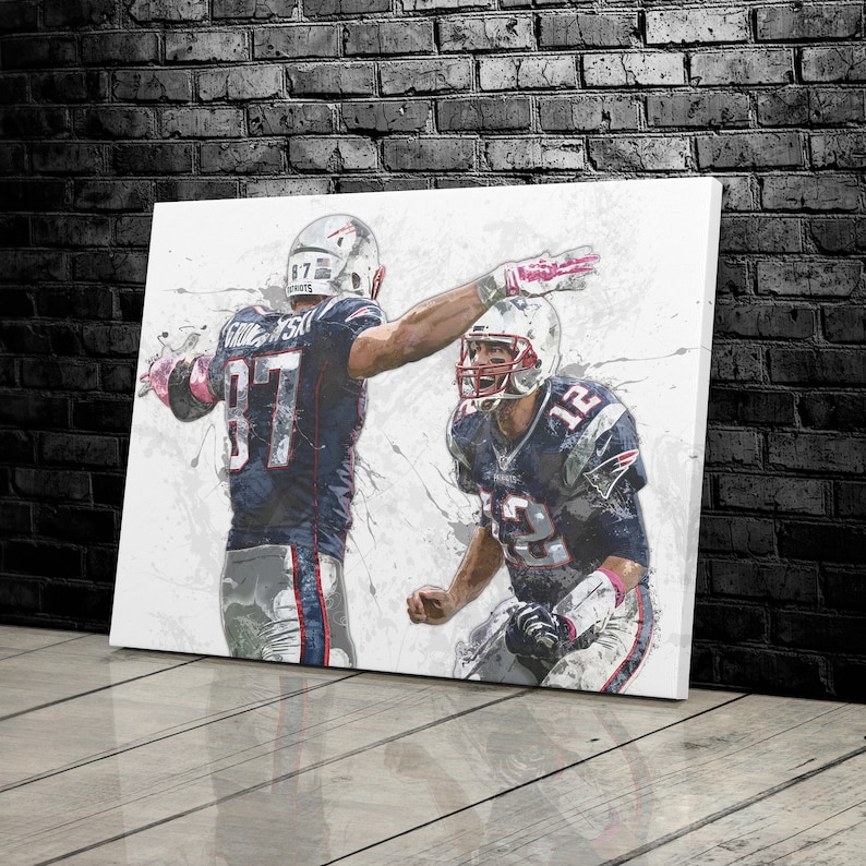 K&ouml;nnte beinhalten: Leinwanddruck mit zwei Footballspielern in Patriots-Trikots. Ein Spieler hebt den Arm zum Jubeln, der andere befindet sich in einer Jubelpose. Das Kunstwerk hat einen Aquarelleffekt mit grauen Spritzern.