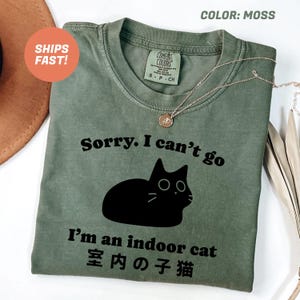 以下が含まれることがあります： 「Sorry. I can't go, I'm an indoor cat」の文字と黒猫のイラストがプリントされたモスグリーンのTシャツ。 「室内猫」の日本語訳もプリントされています。 金色のネックレスがTシャツの上にかけられています。