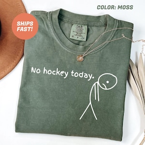 May include: A moss green t-shirt with the text "No hockey today." and a stick figure graphic. The shirt has a crew neck and short sleeves. A gold necklace with a pendant is draped on the shirt.