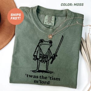 May include: Moss green t-shirt featuring a black illustration of a frog in a kimono holding a sword. The text "'twas the 'tism m'lord" is printed below the frog. A gold necklace is draped across the shirt.