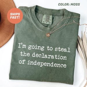 May include: Moss green t-shirt with the text "I'm going to steal the declaration of independence" in white. The shirt has a crew neck and short sleeves. A gold necklace with a pendant is draped on the shirt.