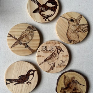 May include: Set of six wooden coasters with detailed pyrography bird designs. Includes a chickadee, sparrow, hummingbird, blue jay, swallow, and hummingbird on a wood slice. Each coaster is round, approximately 4 inches in diameter.