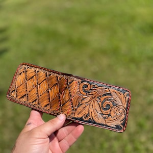 May include: Hand-tooled leather wallet with a basketweave pattern and floral design. The wallet features a brown and black color scheme with red stitching around the edges. The intricate details and craftsmanship are evident in the image.