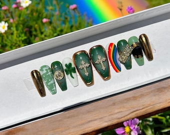 May include: A set of ten green and gold press-on nails with various designs, including shamrocks, crosses, and a rainbow. The nails are displayed in a white box.