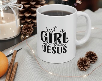 Op de afbeelding: Witte keramische mok met de tekst "just a GIRL who loves JESUS" in zwart schrift. De mok is gevuld met een donkere vloeistof en staat op een lichtgrijze ondergrond, met decoratieve elementen zoals een kaars, dennenappels en lichtslingers.