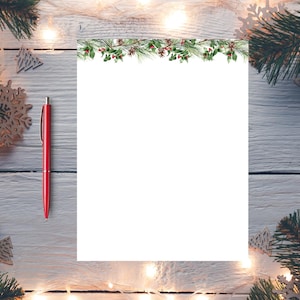 Op de afbeelding: Een wit vel papier met een groene guirlande rand van dennentakken, hulstbesjes en bladeren. Een rode pen ligt op een wit houten oppervlak met een string witte lampjes en houten kerstversieringen.