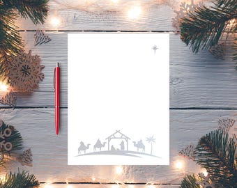 Puede incluir: Una hoja de papel blanca con una ilustración gris de un nacimiento en la parte inferior. Un bolígrafo rojo descansa en el lado izquierdo. El fondo es una superficie de madera blanca decorada con luces navideñas, ramas de pino y adornos de copos de nieve.