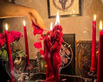 May include: A red wax candle shaped like two figures embracing, set in a dark metal tray. The candle is lit, surrounded by red rose petals and several tall, thin red taper candles. Smoke swirls around the scene, which includes framed artwork.