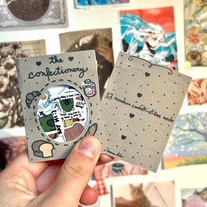 May include: A small brown paper box with a window showing a variety of colorful stickers inside. The box has a heart and the text "the confectionery" written on it. The box also has the text "12 random confetti-stickers inside" written on it.