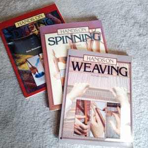 Może przedstawiać: Trzy książki rzemieślnicze zatytułowane "Hands On Dyeing", "Hands On Spinning" i "Hands On Weaving". Książki mają kolorowe okładki ze zdjęciami związanymi z każdym rzemiosłem. Książki są ułożone na szarym dywanie.