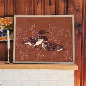 Puede incluir: Una pieza de arte mural rectangular enmarcada que presenta una pintura de dos colimbos con plumaje moteado en marrón oscuro y blanco. Las aves están situadas entre ramas de hojas de color rosa rojizo sobre un fondo de color terracota.