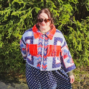 May include: A patchwork jacket with red fringe and a blue and white pattern. The jacket has a red plaid collar and is paired with a navy blue polka dot skirt. The background is a green bush.