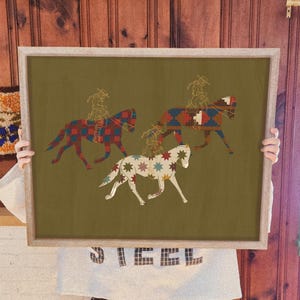 Puede incluir: Una pieza de arte mural rectangular enmarcada con fondo verde oliva que muestra tres caballos y jinetes. Los caballos están decorados con patrones geométricos y estrellas coloridas en rojo, azul y blanco. Los jinetes son líneas doradas.