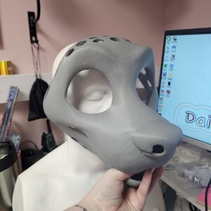 May include: A gray, 3D-printed fursuit head base with large eye openings and a rounded snout. The base is held in front of a white mannequin head. The design includes several ventilation holes.