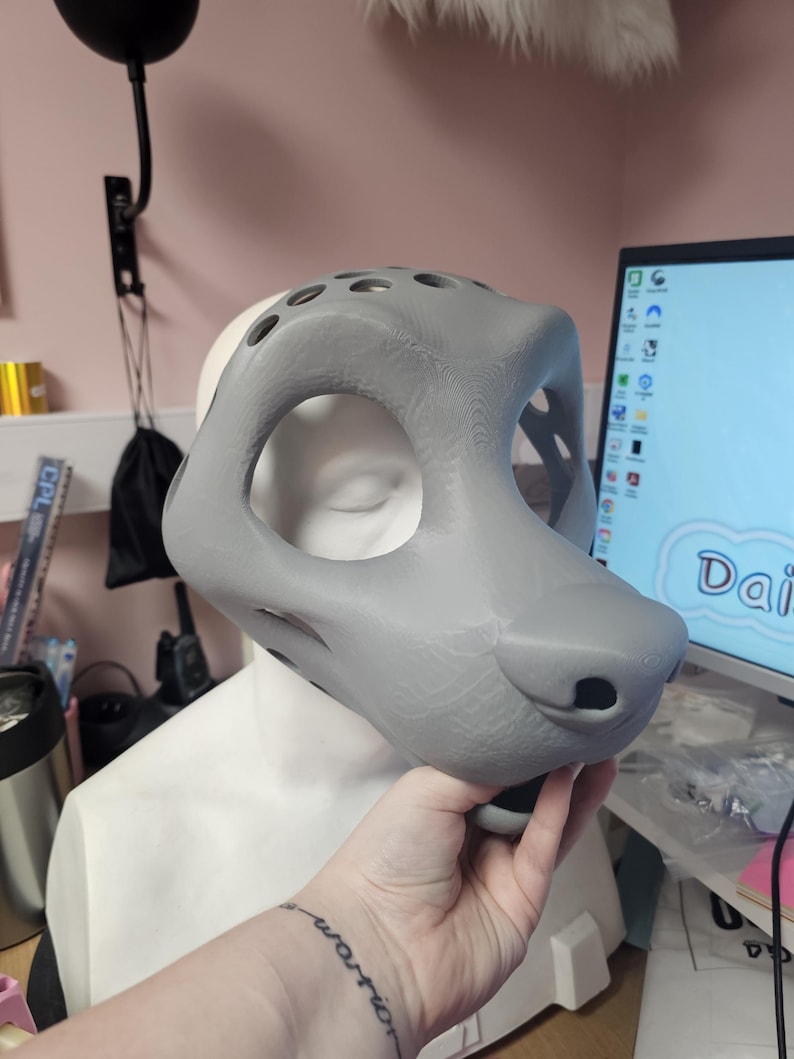 May include: A gray, 3D-printed fursuit head base with large eye openings and a rounded snout. The base is held in front of a white mannequin head. The design includes several ventilation holes.