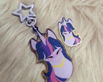 Puede incluir: Un llavero y una pegatina con un unicornio de dibujos animados con pelo morado, rosa y azul. El llavero tiene un cierre en forma de estrella. El unicornio tiene un contorno dorado y un collar amarillo. La pegatina es una versión más pequeña del unicornio.