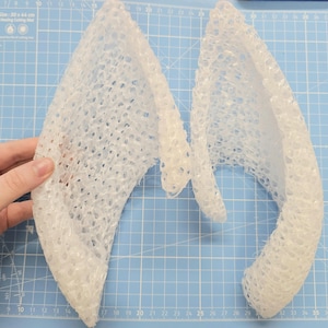 May include: Two white, open-weave, ear-shaped forms, possibly for a costume or craft project, are displayed against a blue grid background. One is held by a hand. The forms have a textured, porous appearance.