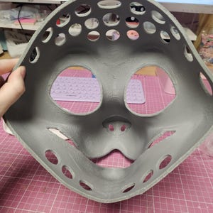May include: A gray, 3D-printed mask with large eye and mouth openings, and numerous circular ventilation holes. The mask has a rounded shape and is held against a pink grid surface.