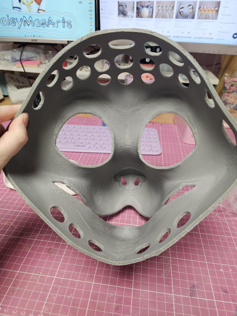 May include: A gray, 3D-printed mask with large eye and mouth openings, and numerous circular ventilation holes. The mask has a rounded shape and is held against a pink grid surface.