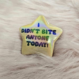Puede incluir: Un pin en forma de estrella con el texto "I DIDN'T BITE ANYONE TODAY!" en azul, verde y amarillo. El pin tiene un acabado holográfico iridiscente y está sobre un fondo rosa suave.