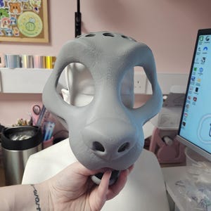 May include: A gray, unfinished fursuit head base, held by a hand. The base has large eye openings, a prominent snout with nostrils, and several ventilation holes on top. The overall shape suggests a canine or feline design.